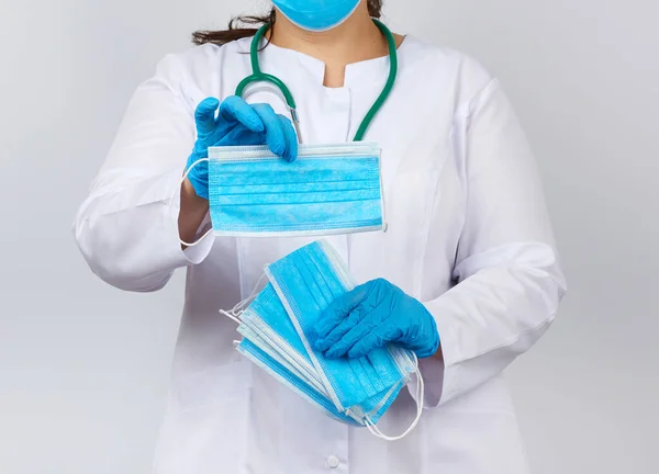 Γυναίκα γιατρός σε ένα λευκό παλτό και μάσκα κρατά μια στοίβα από προστατευτικά — Φωτογραφία Αρχείου