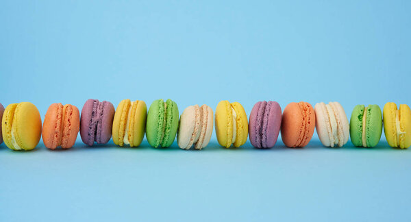 many multi-colored round baked macarons cakes on a light blue background, dessert stands in a row