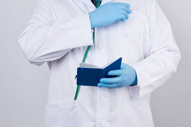Düğmeli beyaz önlüklü erkek doktor, mavi steril eldiven giyiyor, elinde beyaz bir stüdyo arka planında açık mavi bir not defteri tutuyor.