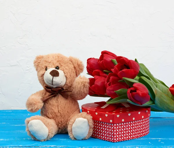 cute brown teddy bear sits on a blue wooden background, bouquet of red tulips, red box, festive backdrop for birthday, Valentine\'s day, anniversary