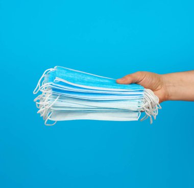 El, mavi arka planda virüslere ve bakterilere karşı tıbbi maske yığını tutuyor. Koruyucu tıbbi araç.