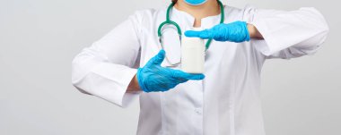 Beyaz önlüklü ve maskeli bir kadın doktor elinde ilaç ve tabletler için beyaz plastik bir kavanoz tutuyor mavi lateks eldivenler, beyaz arka plan