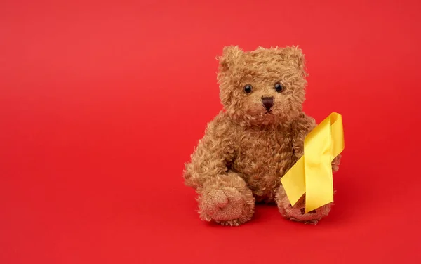 Cute brown teddy bear holds a silk yellow ribbon in the shape of a loop on a red background, concept of the fight against childhood cancer, problem of suicides and their prevention