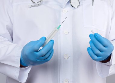 Beyaz önlüklü ve kravatlı bir erkek doktor elinde beyaz arka planda iğnesi olan plastik bir şırınga tutuyor, ellerinde mavi steril tıp eldivenleri, hastalıklara karşı aşı kavramı.