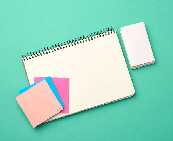 open notebook with blank white sheets, color stickers and rectangular business cards on a green background, top view, flat lay