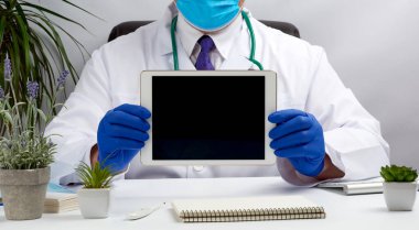 Beyaz bir tıbbi önlükle bir masada oturan ve elinde siyah ekranı olan elektronik bir tablet tutan mavi tıbbi eldiven takan, online tıp konsepti olan