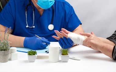 Mavi lateks eldivenli travmatolog doktor masaya oturur ve el yaralanması olan bir hasta resepsiyonu düzenler.