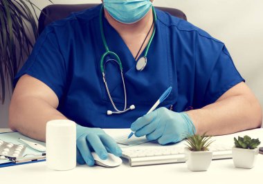 Mavi üniformalı, lateks eldivenli beyaz bir masada oturan erkek doktor, eczane için reçete yazıyor, hasta kabul etmek için tıbbi büro yazıyor.