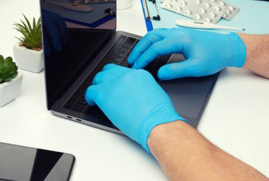 Uzmanlık alanım mavi lateks tıp eldivenleri. Laptop üzerinde çalışıyorum. Uzaktan bakım konsepti, terapist işyeri, üst görüş.