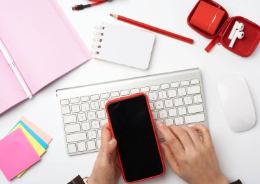 Klavye, bilgisayar faresi, defter ve renk çıkartmaları, üst görünüm, iş yeri, üst görünümü olan beyaz bir masaüstü üzerinde boş bir siyah ekranlı akıllı telefonu tutan eller