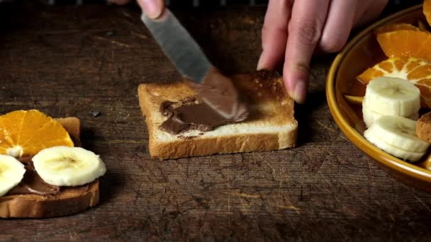 女性の手はトーストパンの正方形の部分に鉄ナイフでチョコレートペーストを粉砕し 上にバナナスライスを産む 茶色の木製のテーブル — ストック動画