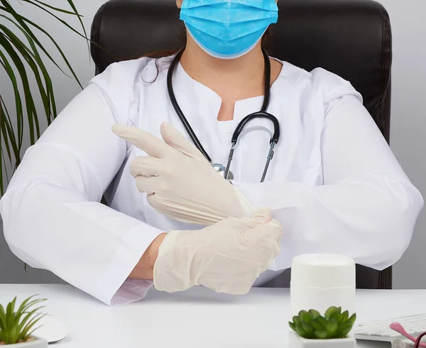 woman doctor in a white uniform, disposable medical mask is sitting in a chair at the table and puts on his hands white sterile latex gloves, doctor office