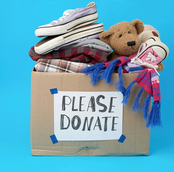 brown cardboard box with a white sheet pasted and the inscription please donate on a blue background, inside clothes, toys, help concept