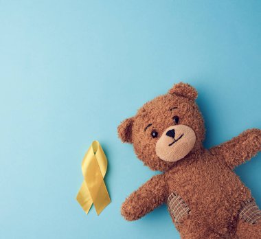 Kahverengi ayıcığın pençesinde mavi arka planda katlanmış sarı bir kurdele var. Çocukluk kanseriyle mücadele kavramı. İntihar ve onların önlenmesi sorunu. Boşluğu kopyala