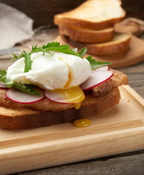 sandwich on toasted white slice of bread with poached eggs, green leaves of arugula and radish, morning breakfast on a brown wooden board