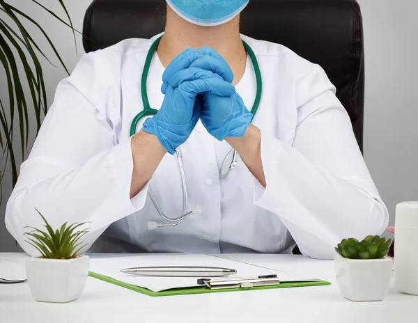 doctor in a white coat and blue latex gloves sits at a white work table in his office, concept of patient reception in the clinic, hands joined in front of the chest, focused pose