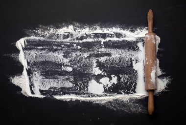 Siyah bir masaya saçılmış beyaz buğday unu ve siyah bir masanın üzerinde ahşap bir oklava.