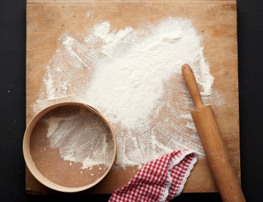 Siyah bir masaya saçılmış beyaz buğday unu ve siyah bir masaya saçılmış ahşap bir merdane, yuvarlak elek, üst görünüm