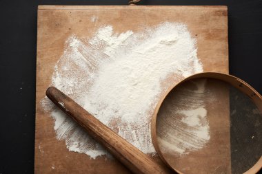 Siyah bir masaya saçılmış beyaz buğday unu ve siyah bir masaya saçılmış ahşap bir merdane, yuvarlak elek, üst görünüm