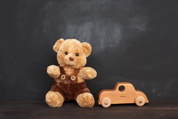 brown teddy bear sits on a brown wooden table and wooden car, behind an empty black chalk board, place for an inscription, back to school