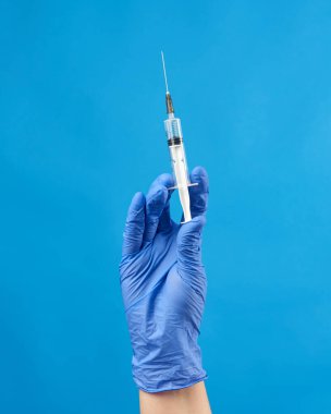 Mavi steril lastik eldivenli kadın eli mavi arka planda beyaz bir şırınga tutuyor.