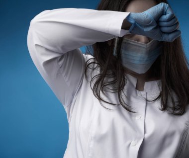 Beyaz önlüklü, mavi lateks eldivenli ve yüzünde koruyucu maske olan genç bir kadın doktor mavi arka planda duruyor. Alnına bastırılmış, yorgunluk ve umutsuzluk kavramı..