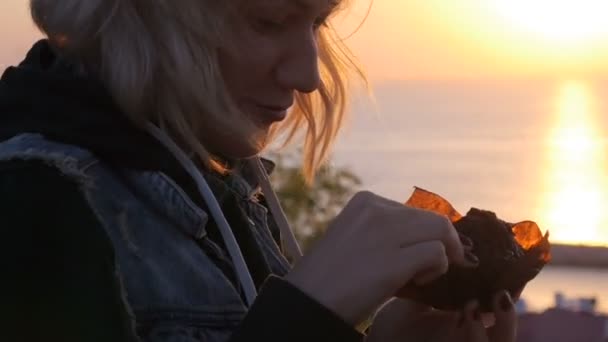Улыбка девушки едят кексы наслаждаясь моментом, когда восход солнца отражается на море — стоковое видео