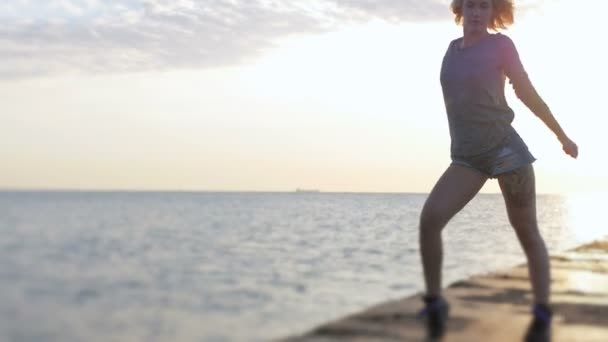 Блондинка в джинсовых шортах танцует счастливо возле пляжа на прекрасном восходе солнца — стоковое видео