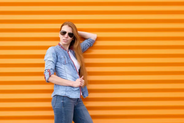 Рыжая девушка в джинсовой рубашке с солнцезащитными очками возле оранжевой стены — стоковое фото
