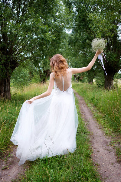 Bride in white llight wedding dress with bridal bouquet walking in lane of trees and dancing, view from behind