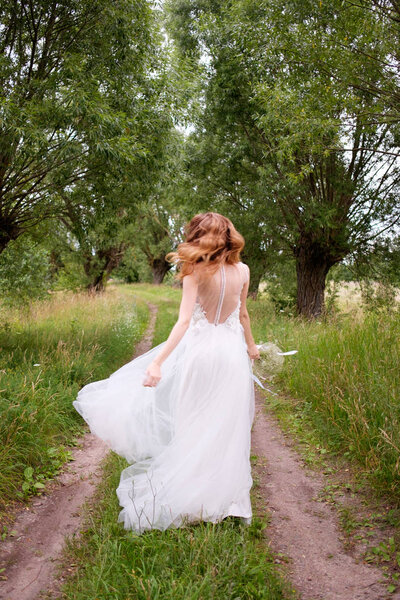 Bride in white llight wedding dress with bridal bouquet running away in lane of trees, view from behind