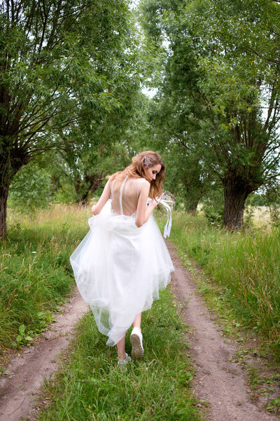 Bride in white llight wedding dress with bridal bouquet running away in lane of trees, view from behind