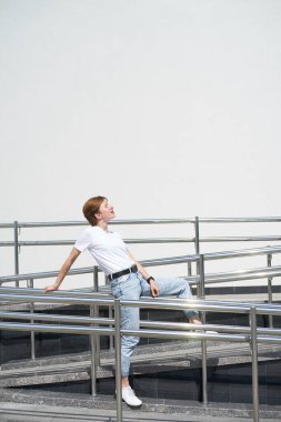 Kısa saçlı, beyaz saçlı, genç bir kadın sıcak yaz gününde şehir merkezinde korkuluklarda oturuyor ve gülümsüyor.