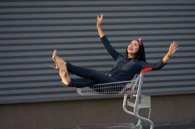 Yeni moda koleksiyon. Komik, mutlu, şık esmer, uzun saçlı, modaya uygun kot pantolon giyen, alışveriş arabasıyla gezen kız. Gri kentsel duvar arka planında çekilen.                               