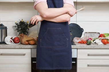 Önlük giyen beyaz adam mutfakta duruyor ve elinde sebzeler ve ahşap kaşıkla yemek pişirmeye hazırlanıyor.