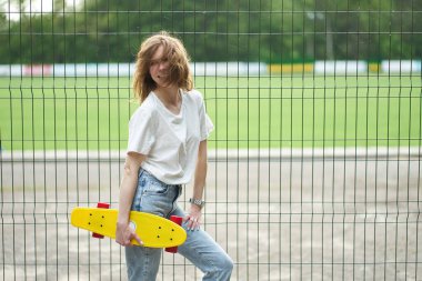 Gülümseyen kızıl saçlı beyaz kız sarı kaykay tutuyor ve çitin yanında duruyor.