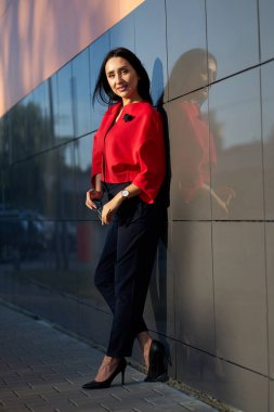 Bahar modern koleksiyonu. Elinde şık güneş gözlüğü olan, şık kırmızı ceketli ve gri arka planda mavi pantolonlu çekici bir kız. Açık havada portre.                              