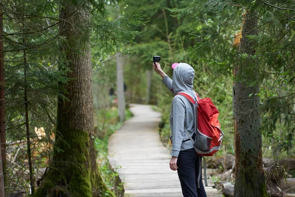 旅行だ 若いです女の子旅行者とともにオレンジバックパック歩くと上の木製の橋で自然保護区と撮影ビデオとともにアクションカメラ — ストック写真