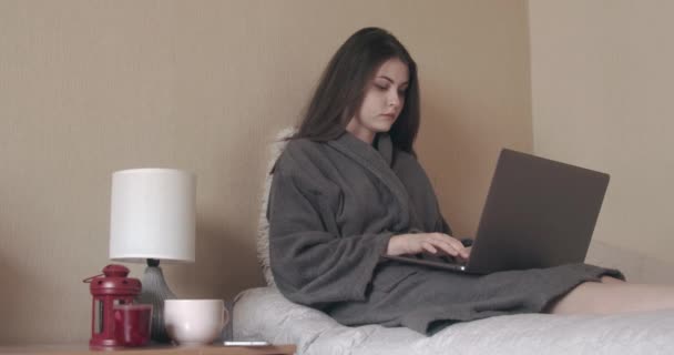 Mooie jonge vrouw zit op bed en werkt op laptop. — Stockvideo