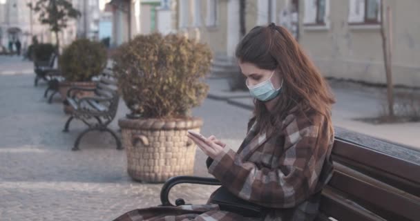 Fiatal nő védő maszkban mobiltelefont használ, miközben a padon ül. — Stock videók