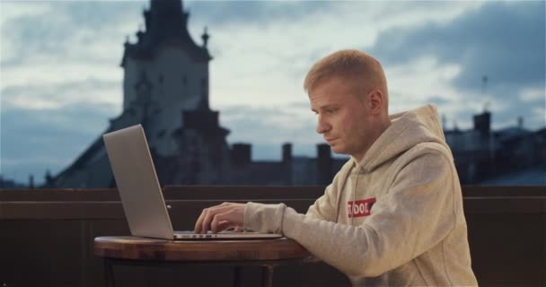 Um jovem sentado no telhado com um laptop com uma vista da torre da cidade velha no fundo — Vídeo de Stock