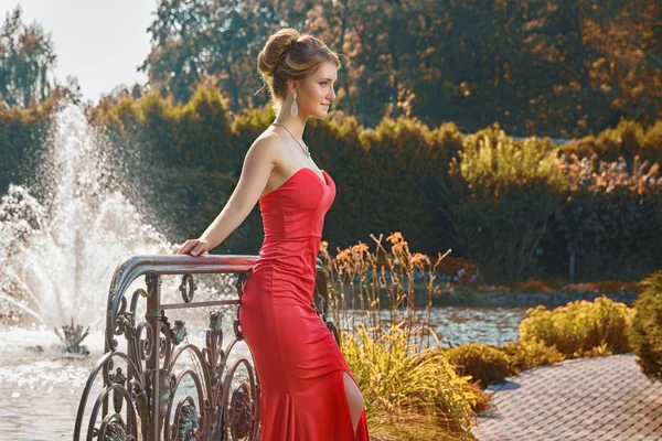 Elegant young woman in red dress standing in front of fountain. — Stock Photo, Image