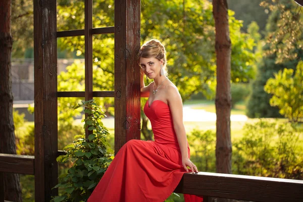 Ελκυστική νεαρή λεπτή γυναίκα στο πάρκο, κυρία με κόκκινο φόρεμα και τα ψηλά τακούνια έχει διασκέδαση, κάθεται — Φωτογραφία Αρχείου