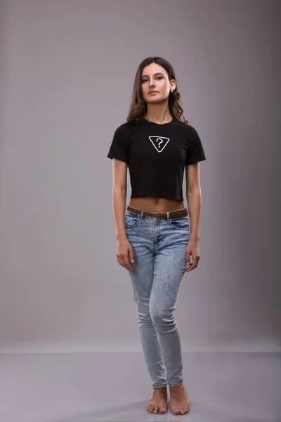 广告和 t 恤的设计理念。时尚的女人，身穿黑色 t 恤与题字问号和蓝色牛仔裤站在反对与副本空间灰色工作室的墙上的肖像 — 图库照片
