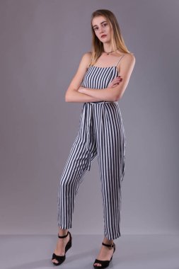 Confident attractive slender young woman wearing striped overall standing with folded arms looking thoughtfully at the camera over gray studio background clipart