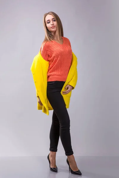 Elegancia chica está posando en el estudio sobre el fondo gris. Lleva pantalones negros, jersey naranja y abrigo amarillo. . —  Fotos de Stock