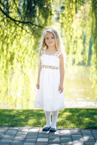 Sevimli küçük sarışın kız arkadan aydınlatmalı su ve çim ağaç arka planı olduğunu — Stok fotoğraf