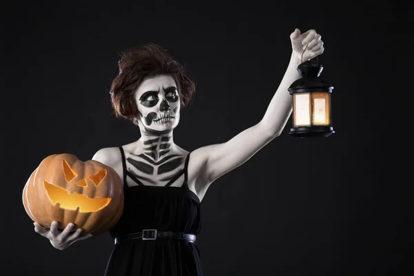 Glad halloween. Porträtt av svart häxa med skrämmande smink över svart bakgrund. Hålla pumpa och lampa i händer — Stockfoto