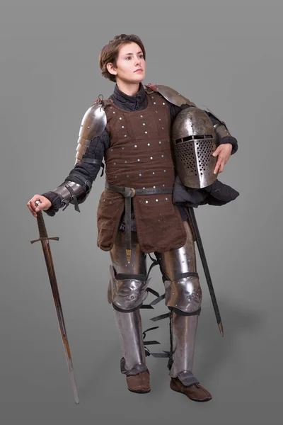 Молодая женщина в рыцарской броне держит шлем в одной руке и меч на плече на сером фоне — стоковое фото