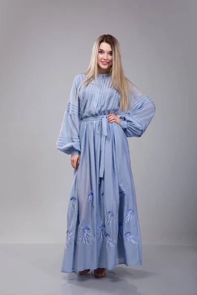 Mooie jonge vrouw dresseed in lange blauwe geborduurde jurk op grijs studio achtergrond — Stockfoto